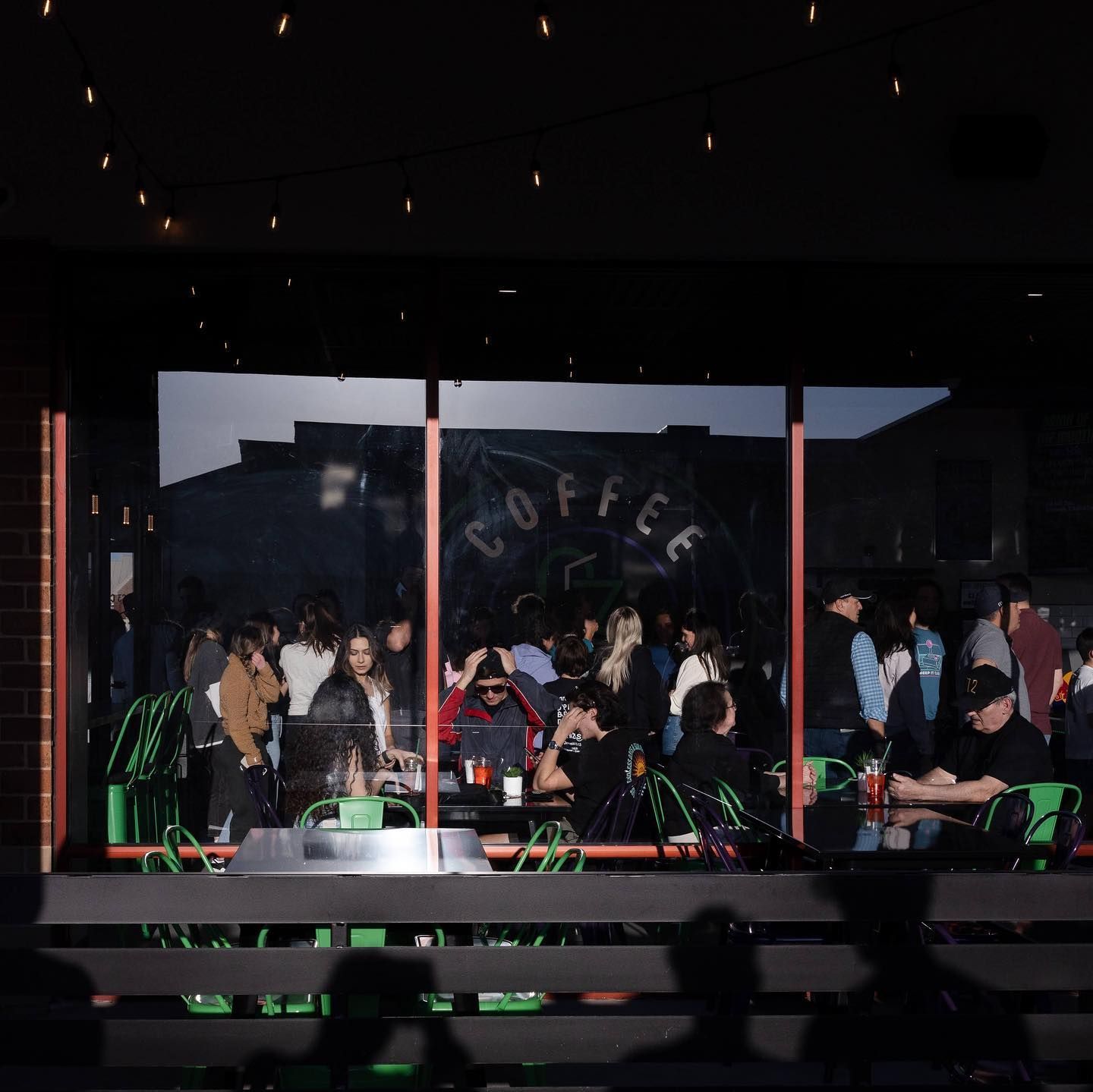 People at outdoor cafe tables in front of a coffee shop window; green chairs, dim lighting.