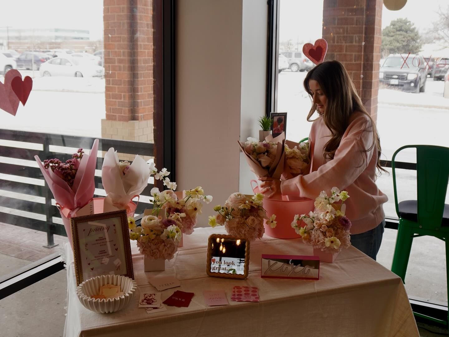 Woman arranging flower bouquets at a table near a window, Valentine's Day decor, pink theme.