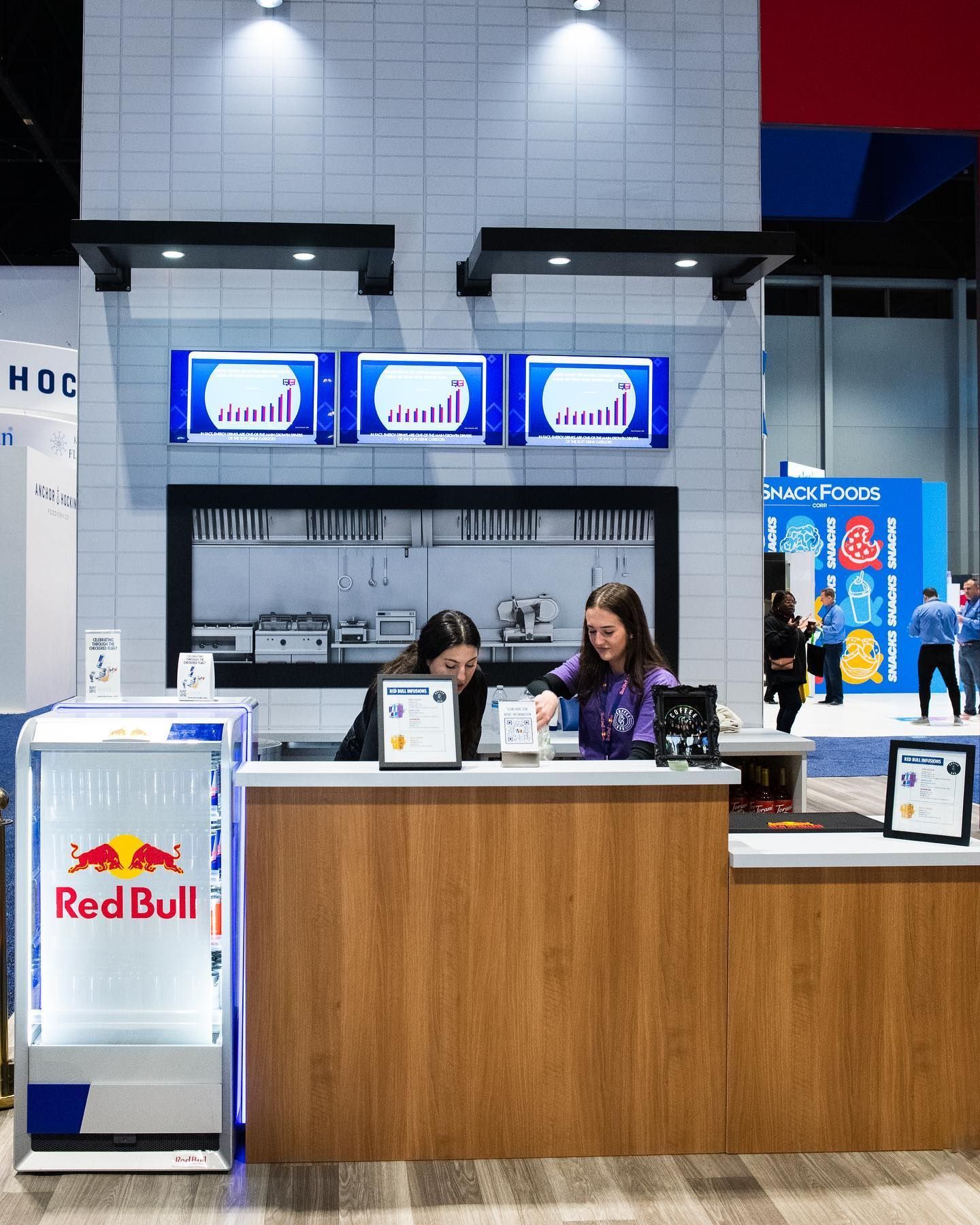Two people at a wooden counter in a booth with a Red Bull fridge. Monitors show graphs. White tile background.