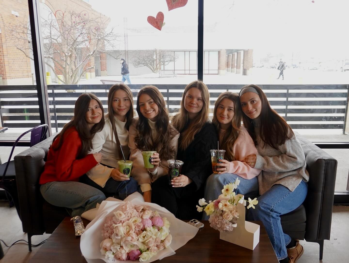 Six smiling people seated on a couch with bouquets, drinks. Indoor setting with a window view.