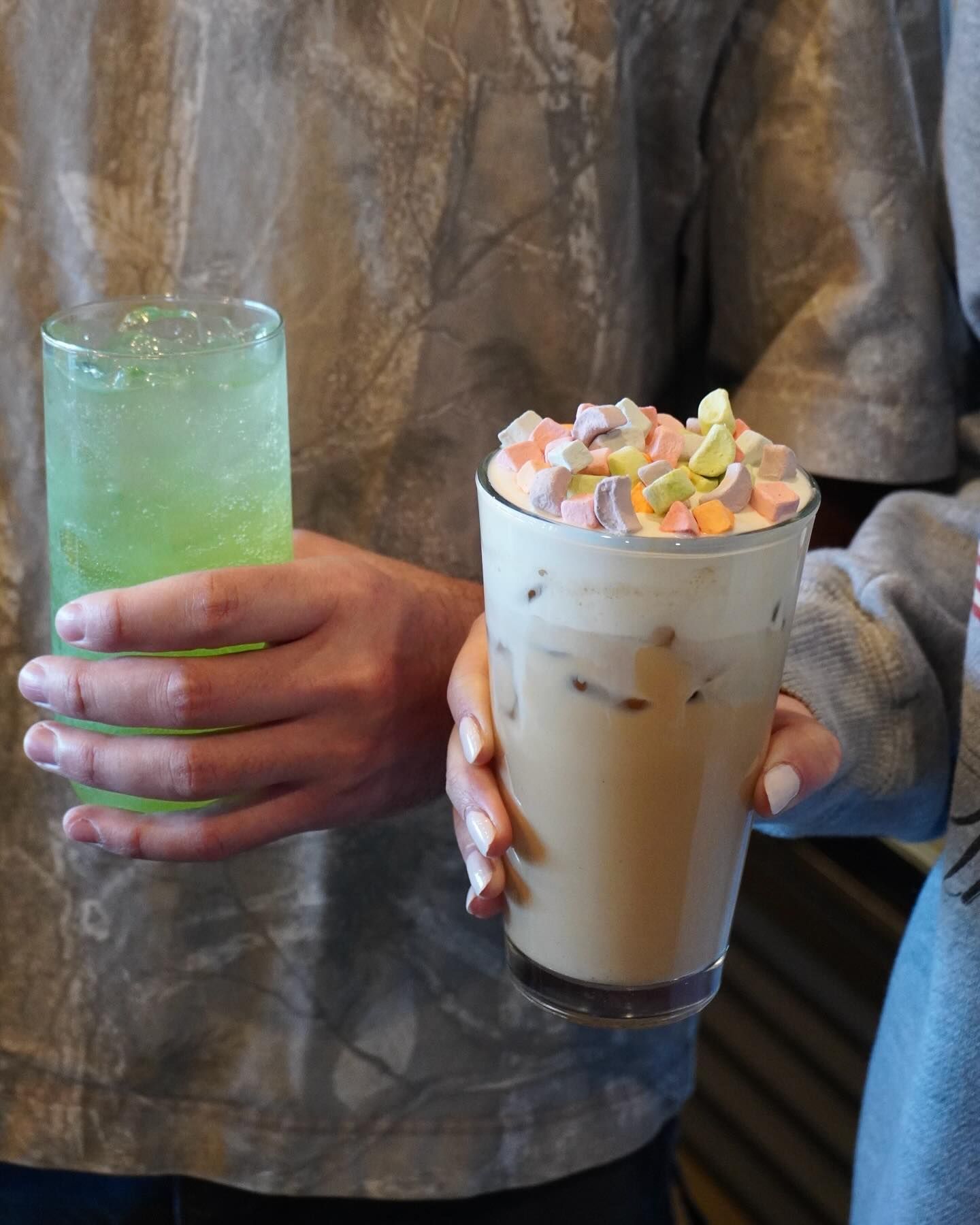 Two drinks: green soda and iced coffee topped with marshmallows, held by hands.