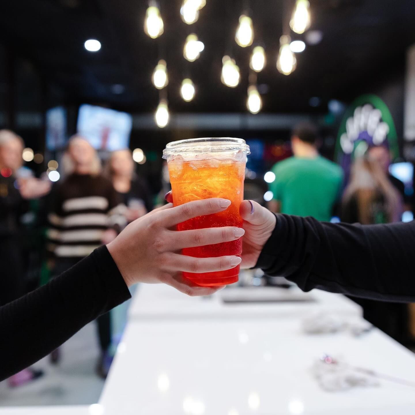 Two hands clink a colorful, layered drink. Blurred bar scene in the background with overhead lights.