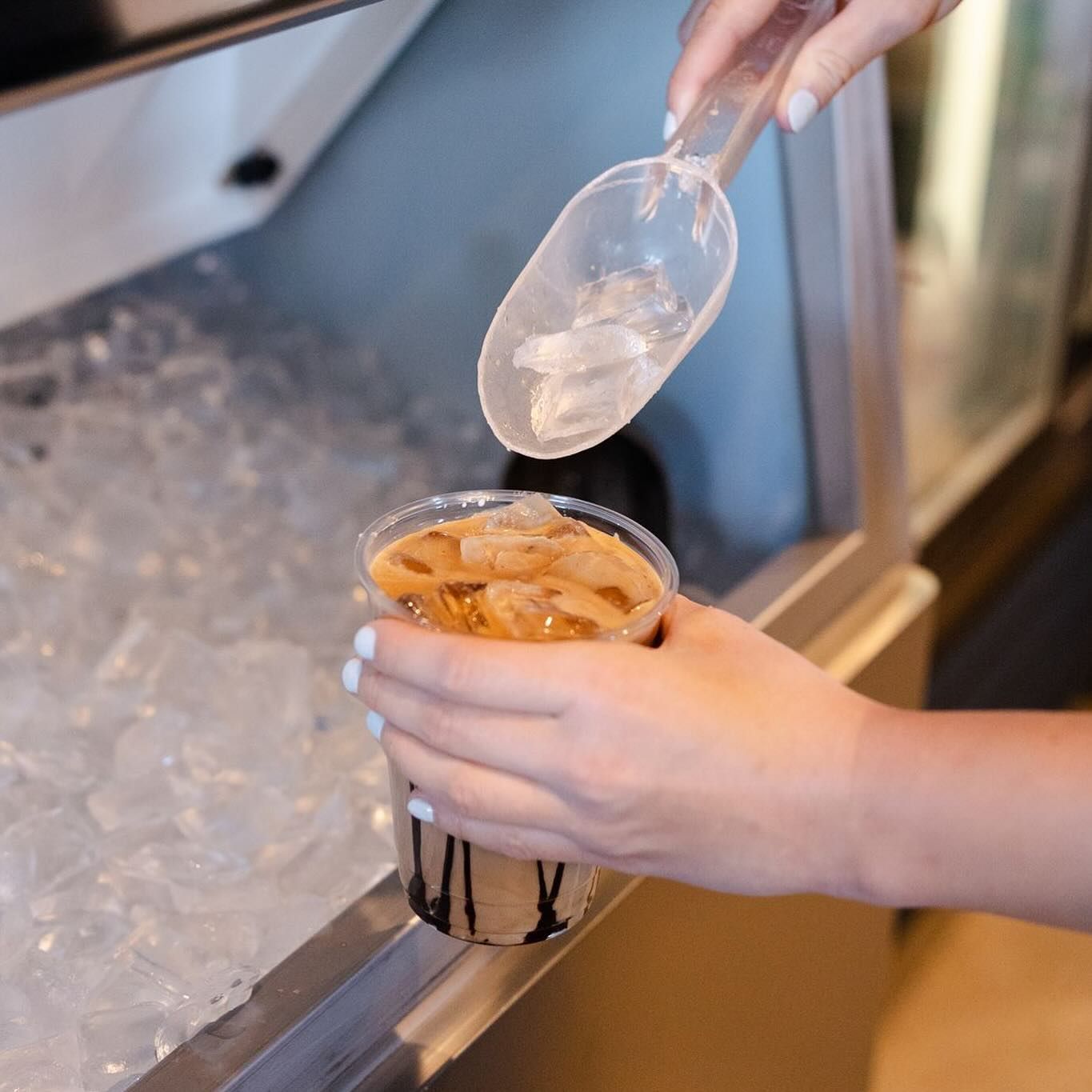 Person adding ice cubes to iced coffee in a clear plastic cup, using a clear plastic scoop.