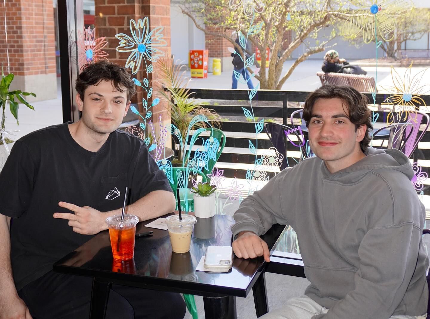 Two people sitting at an outdoor cafe table. One person giving peace sign. Drinks on the table.