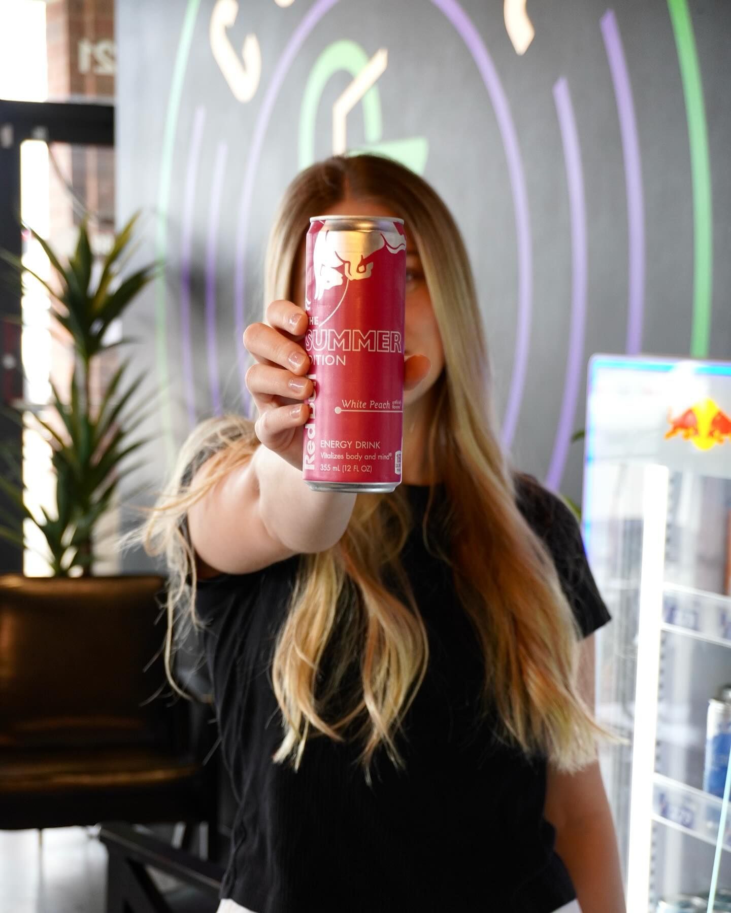 Woman holding a Red Bull can in front of her face; colorful abstract background.