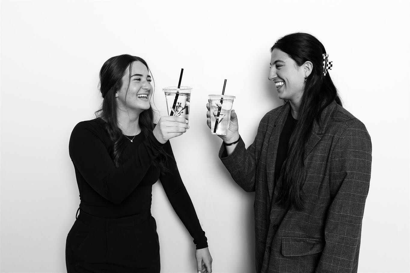 Two people, smiling and toasting iced drinks with straws. Black and white photo against a plain backdrop.