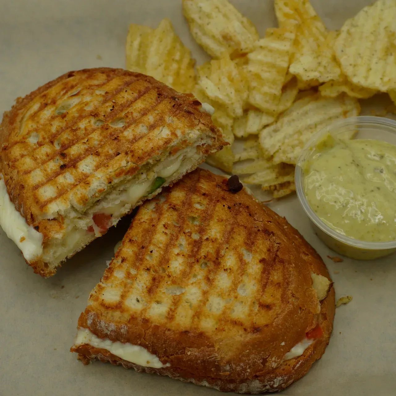 A grilled panini cut in half, served with a pile of ripple-cut potato chips and a small container of dipping sauce.