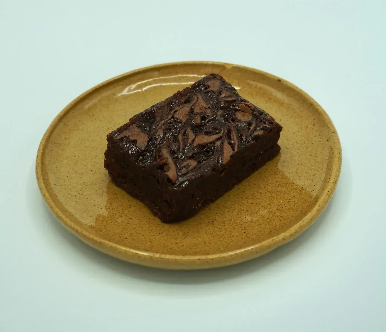 A square, marbled chocolate brownie sits centered on a round, speckled tan plate against a plain white background.