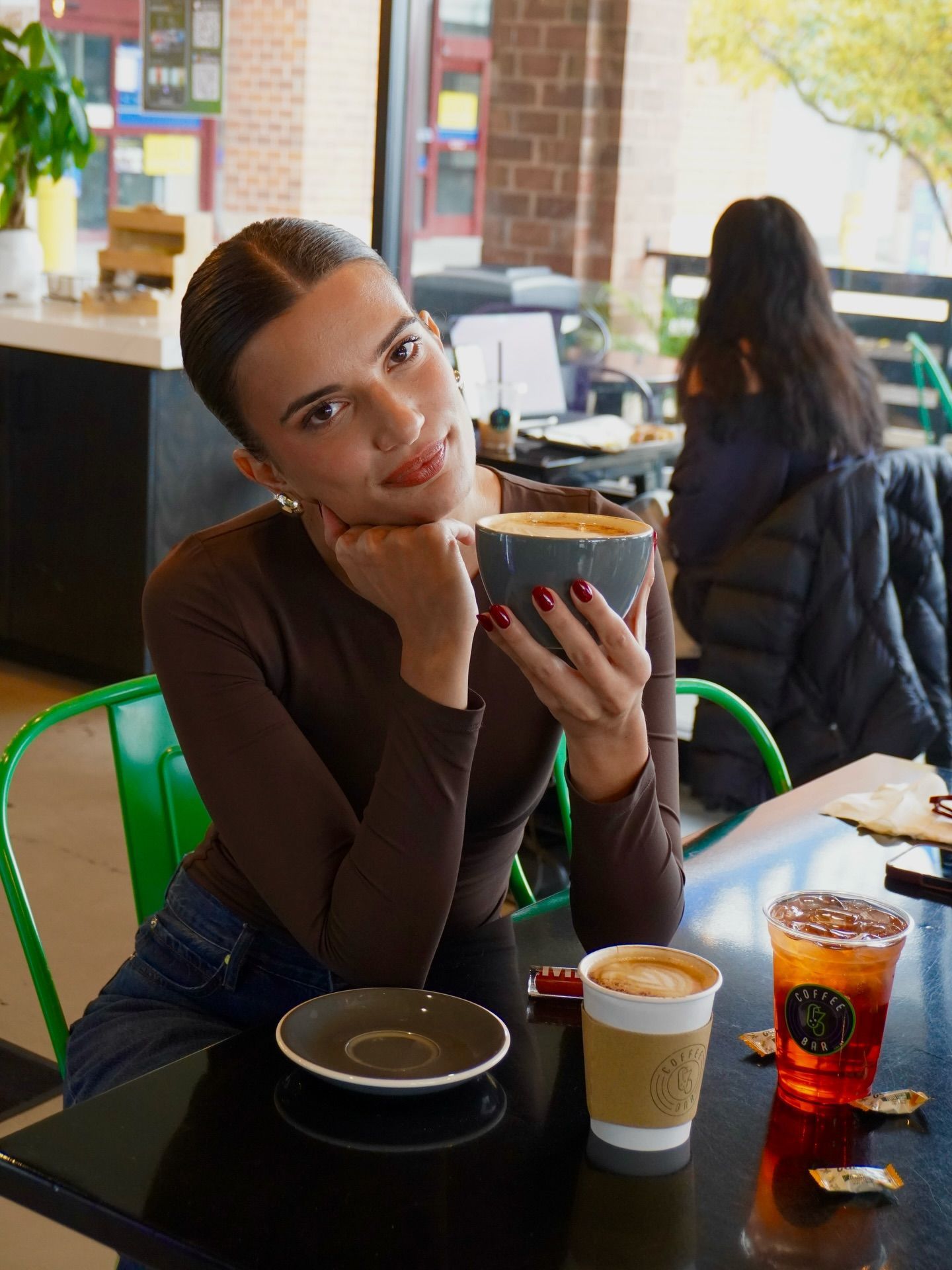 Woman smiles, holding a coffee cup in a cafe setting, other drinks on the table.
