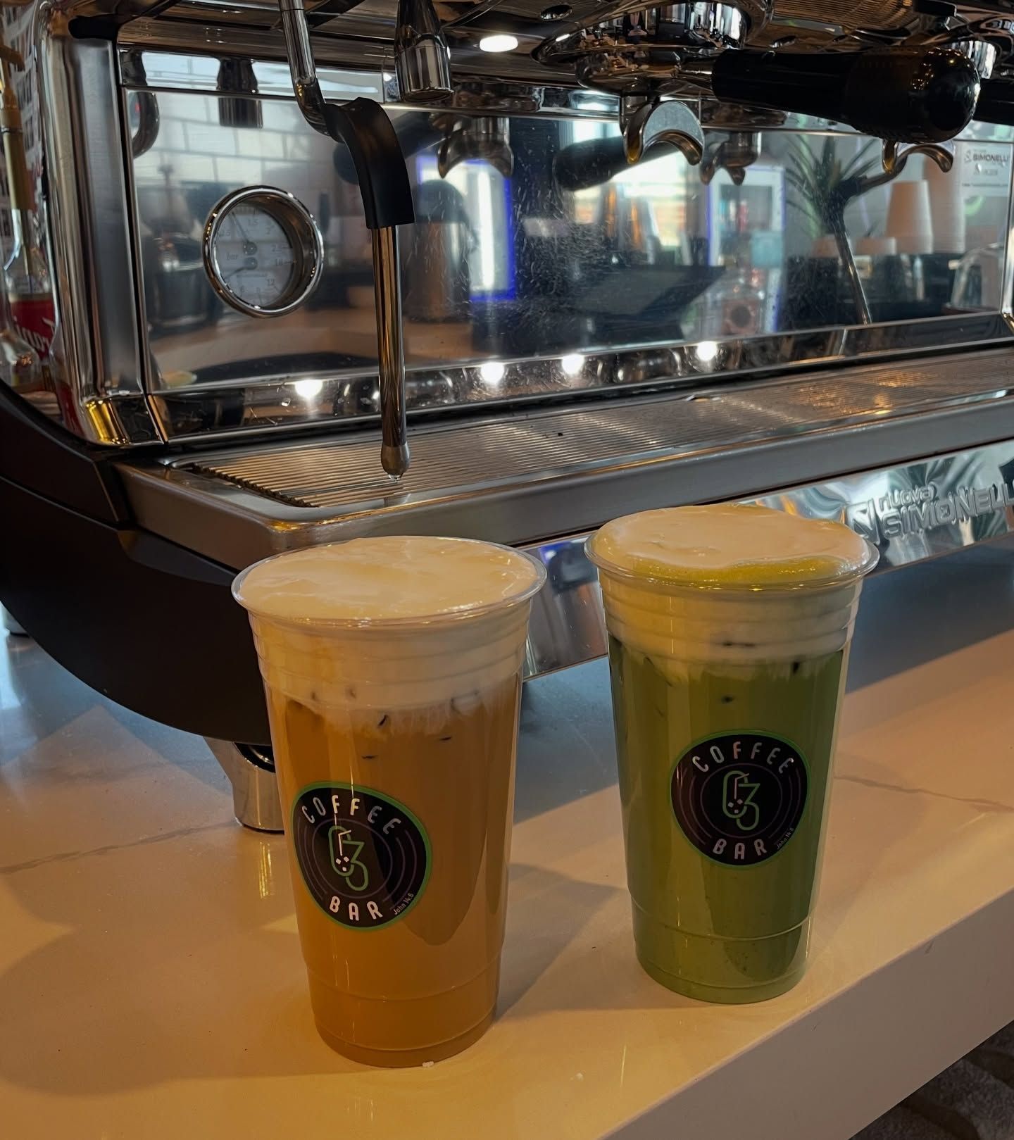 Two iced drinks with foam, orange and green, in front of an espresso machine at a coffee shop.