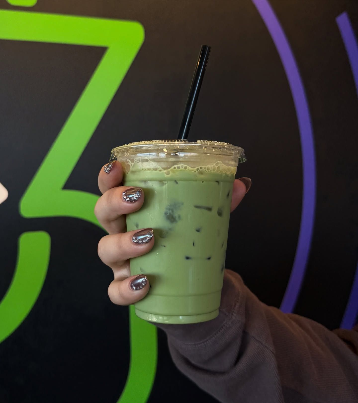 Hand holding a clear cup of iced matcha latte with a black straw against a colorful background.