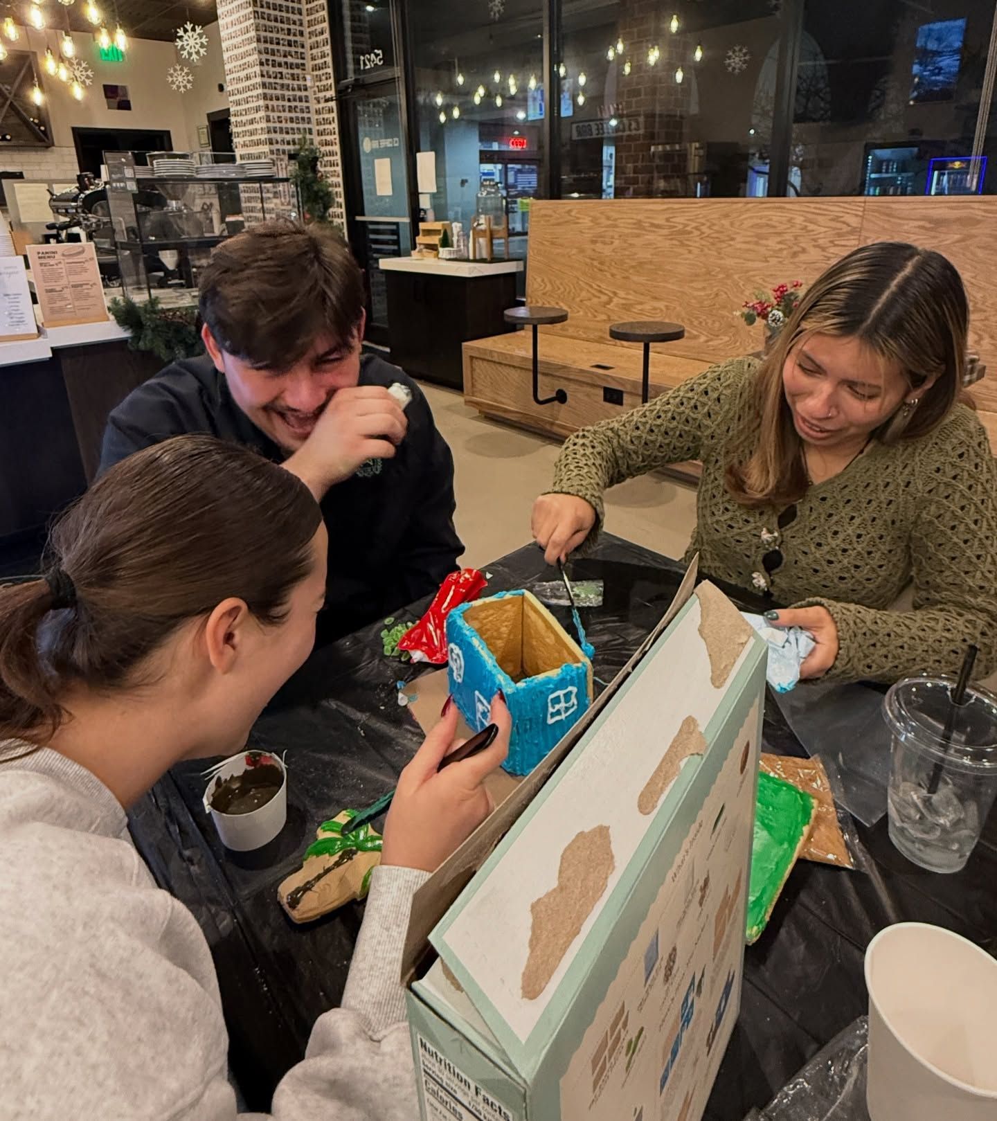 Three people painting at a table indoors: A blue birdhouse, and other items are spread around on a black surface.