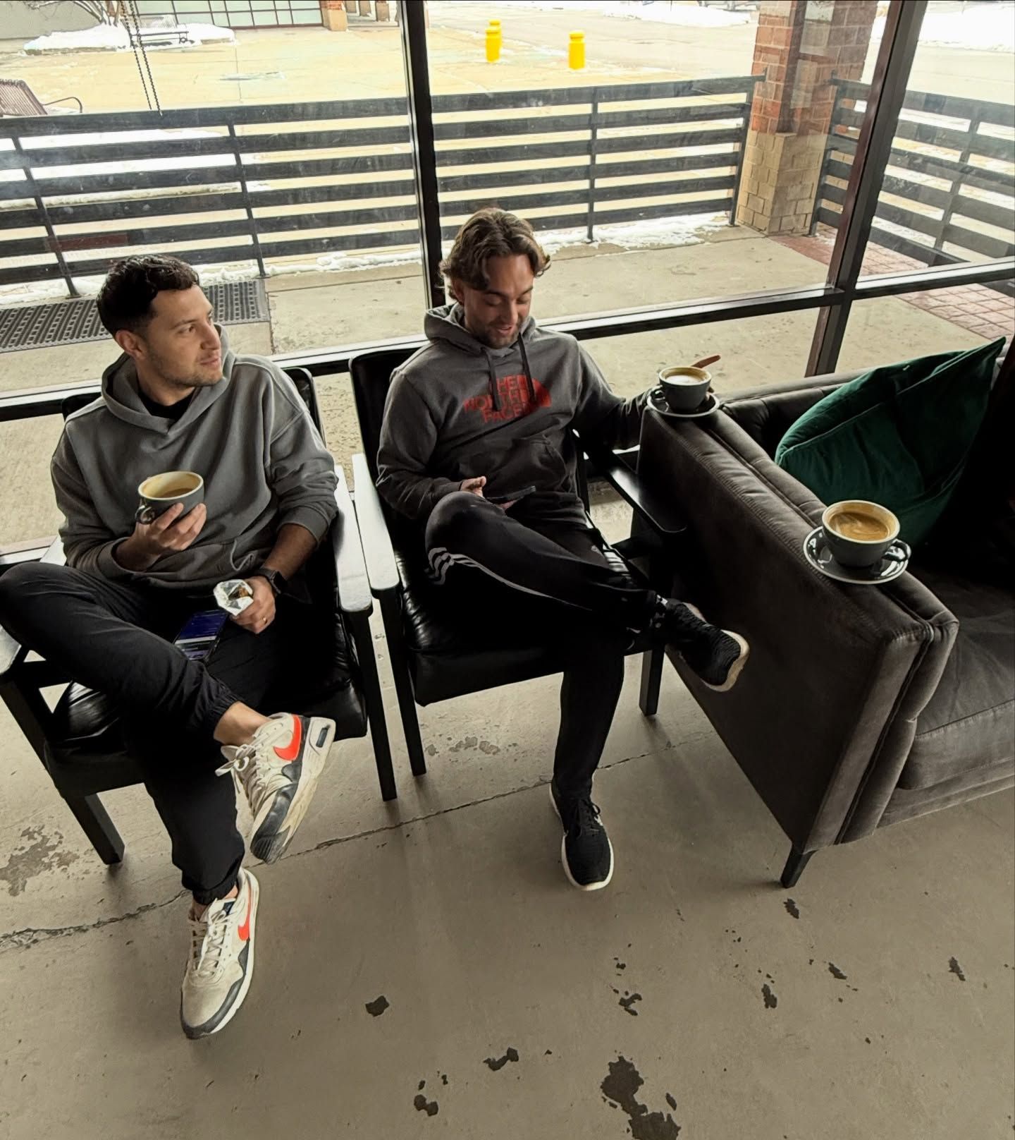 Two men in hoodies sit, drinking coffee. One looks left, the other at his phone. Outdoor cafe setting.