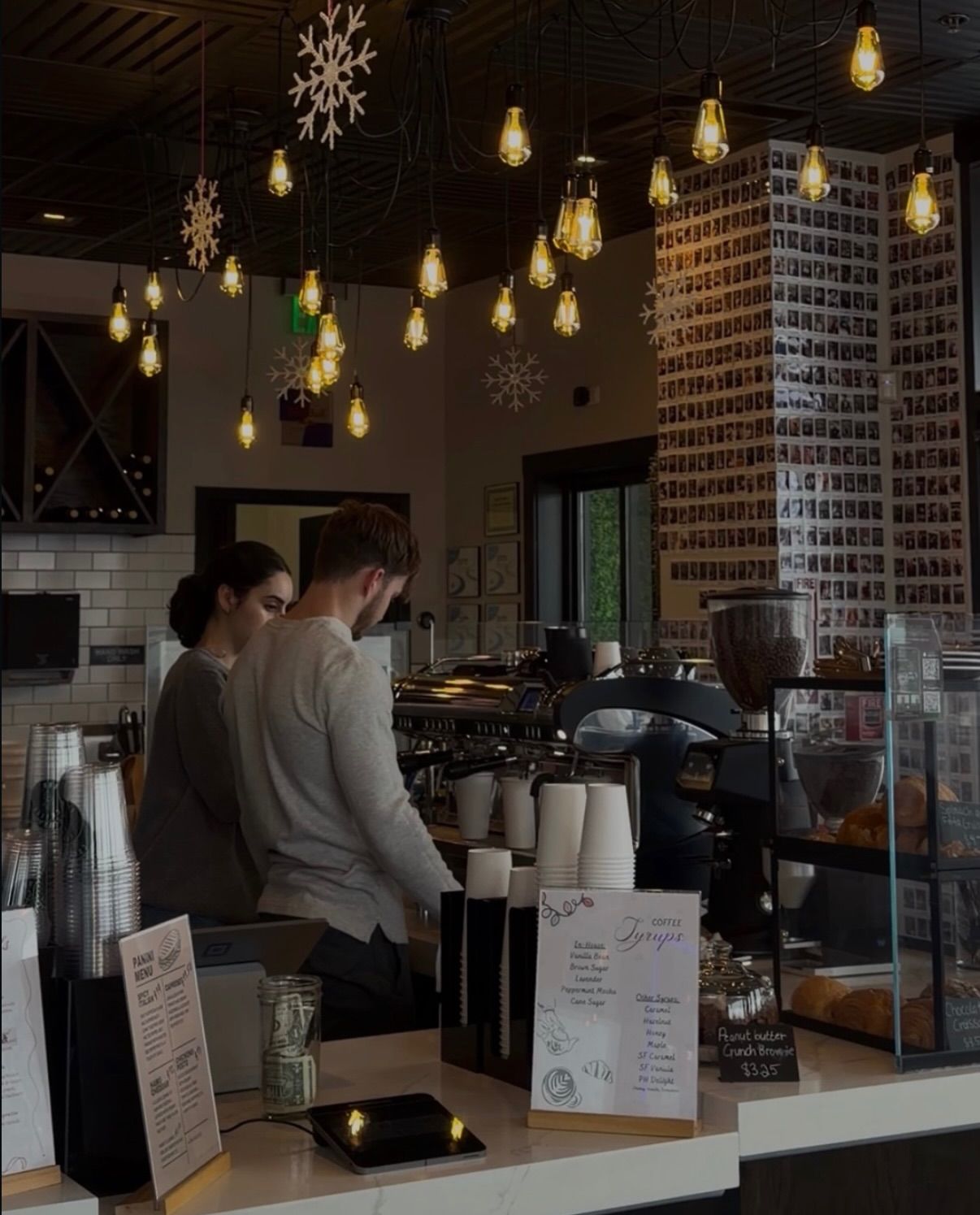 Two people behind a coffee shop counter, preparing drinks. Overhead lights and snowflakes decorate the ceiling.