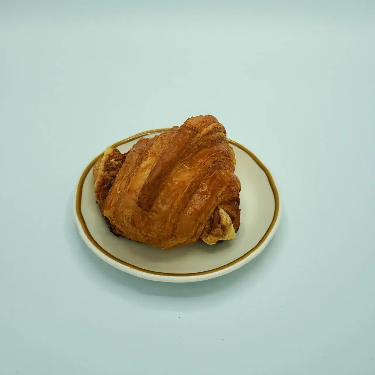 Croissant pastry on a small white plate with a brown rim, set against a light blue background.