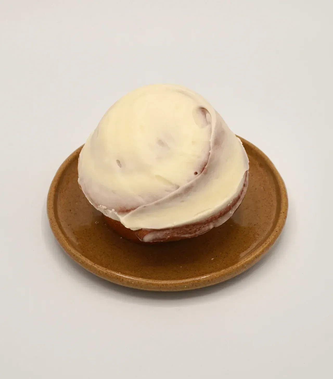 A single frosted cinnamon roll sits centered on a small, glossy brown plate against a plain white background.