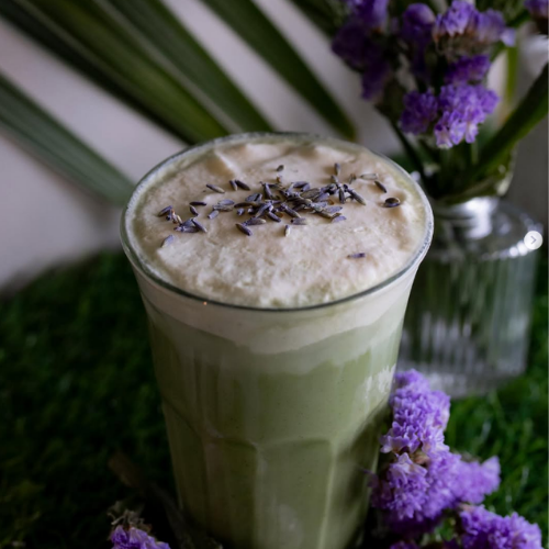Green matcha latte with frothy top, lavender garnish, purple flowers, and a clear glass.