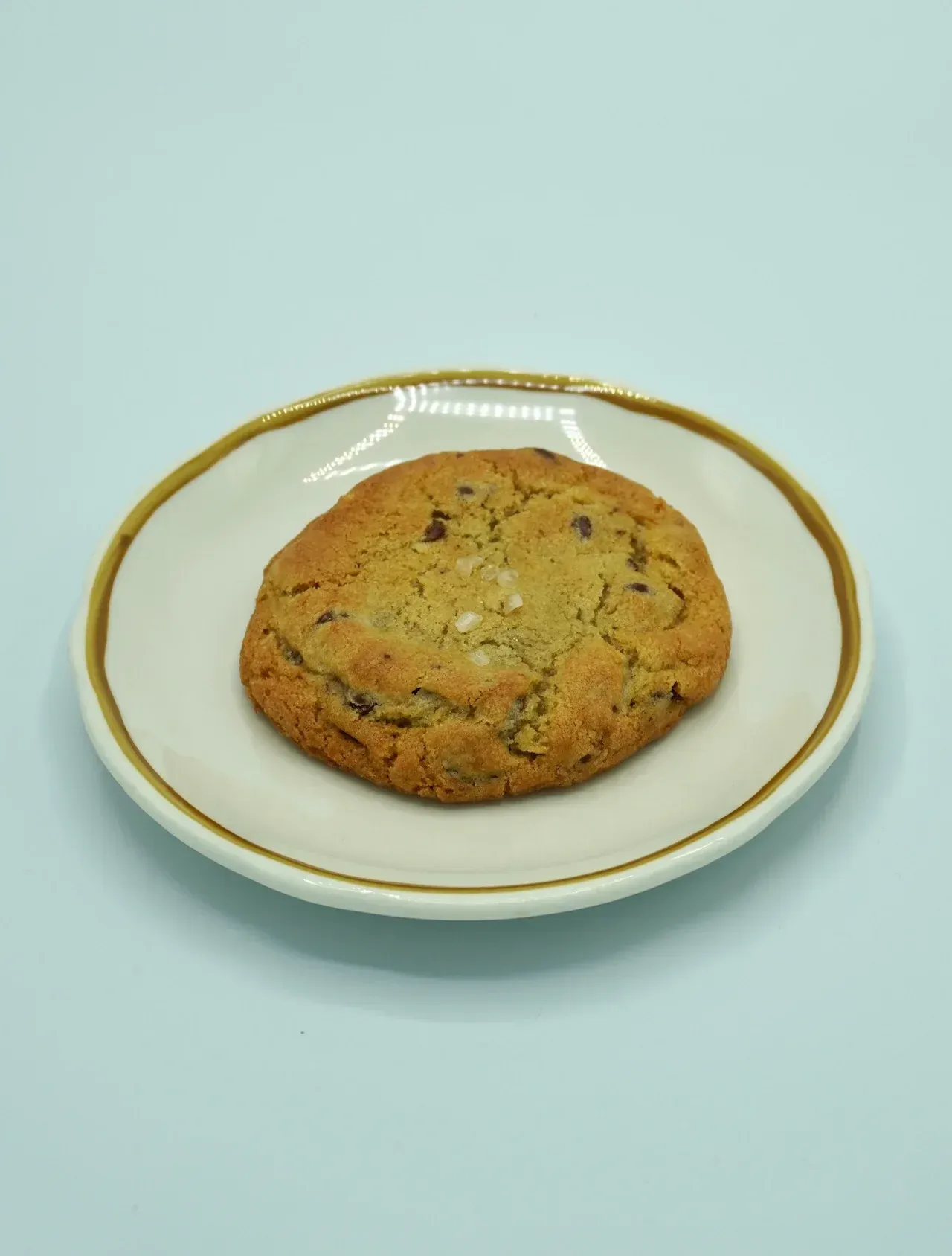 A single chocolate chip cookie centered on a white, gold-rimmed plate against a light blue background.