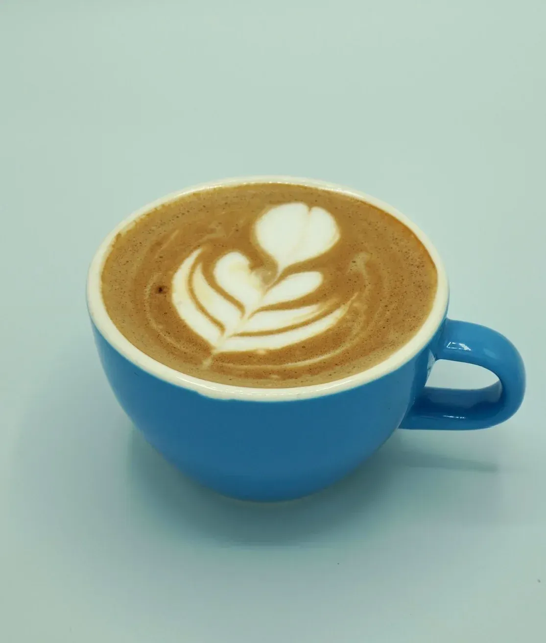 Blue cup of latte with heart and tulip latte art, on a light blue surface.