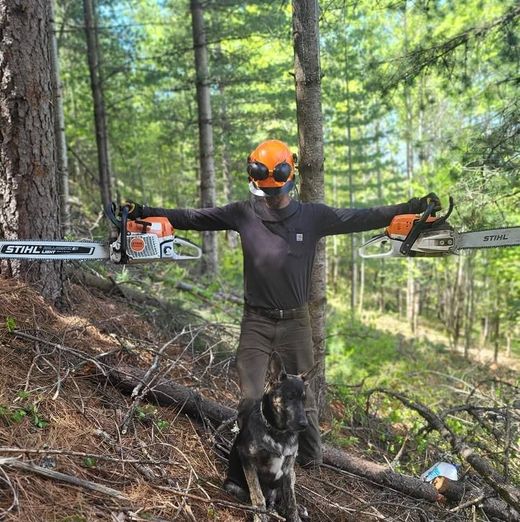 A man and a dog are standing in the woods holding chainsaws.