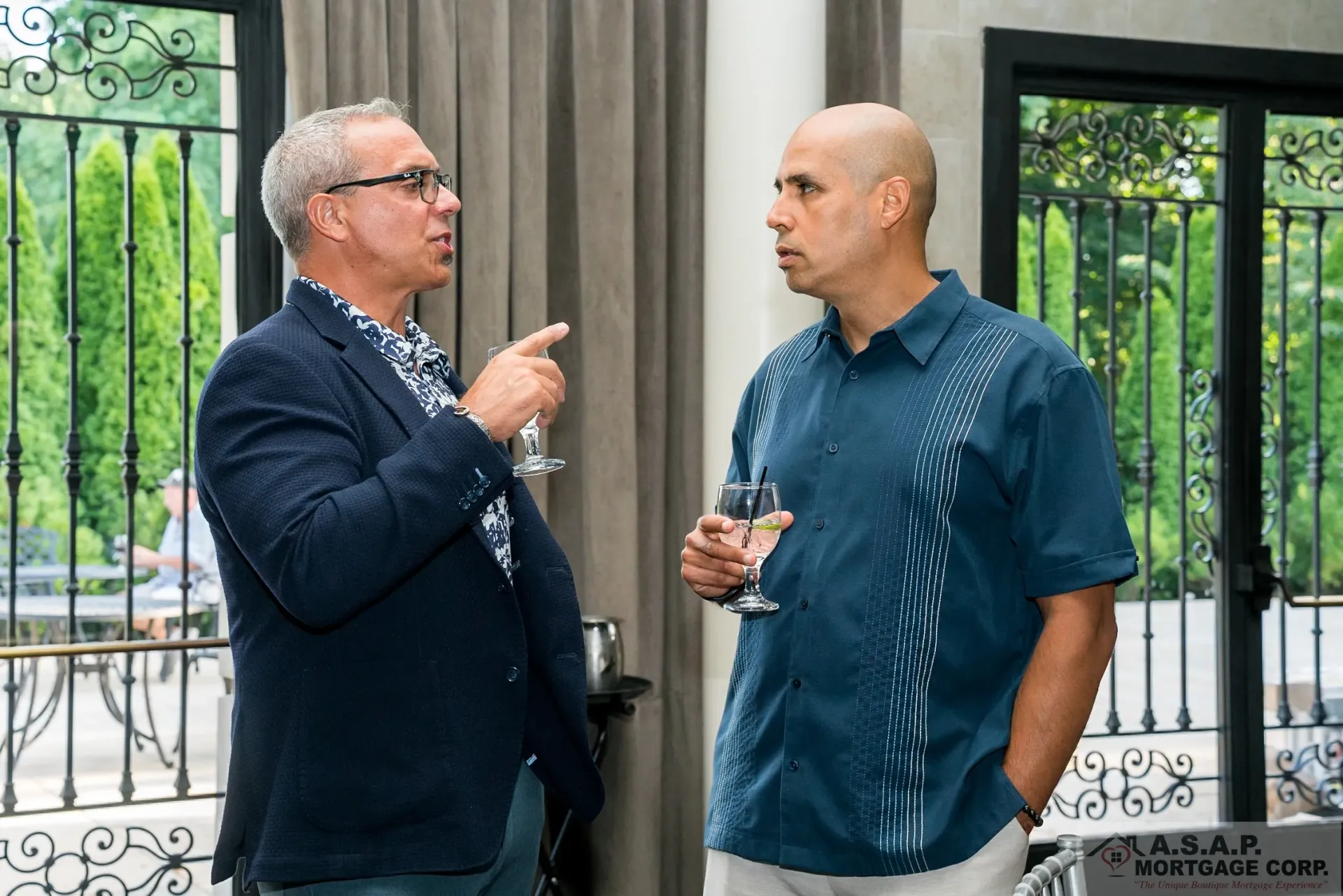 Two men are talking to each other in a room while holding wine glasses.