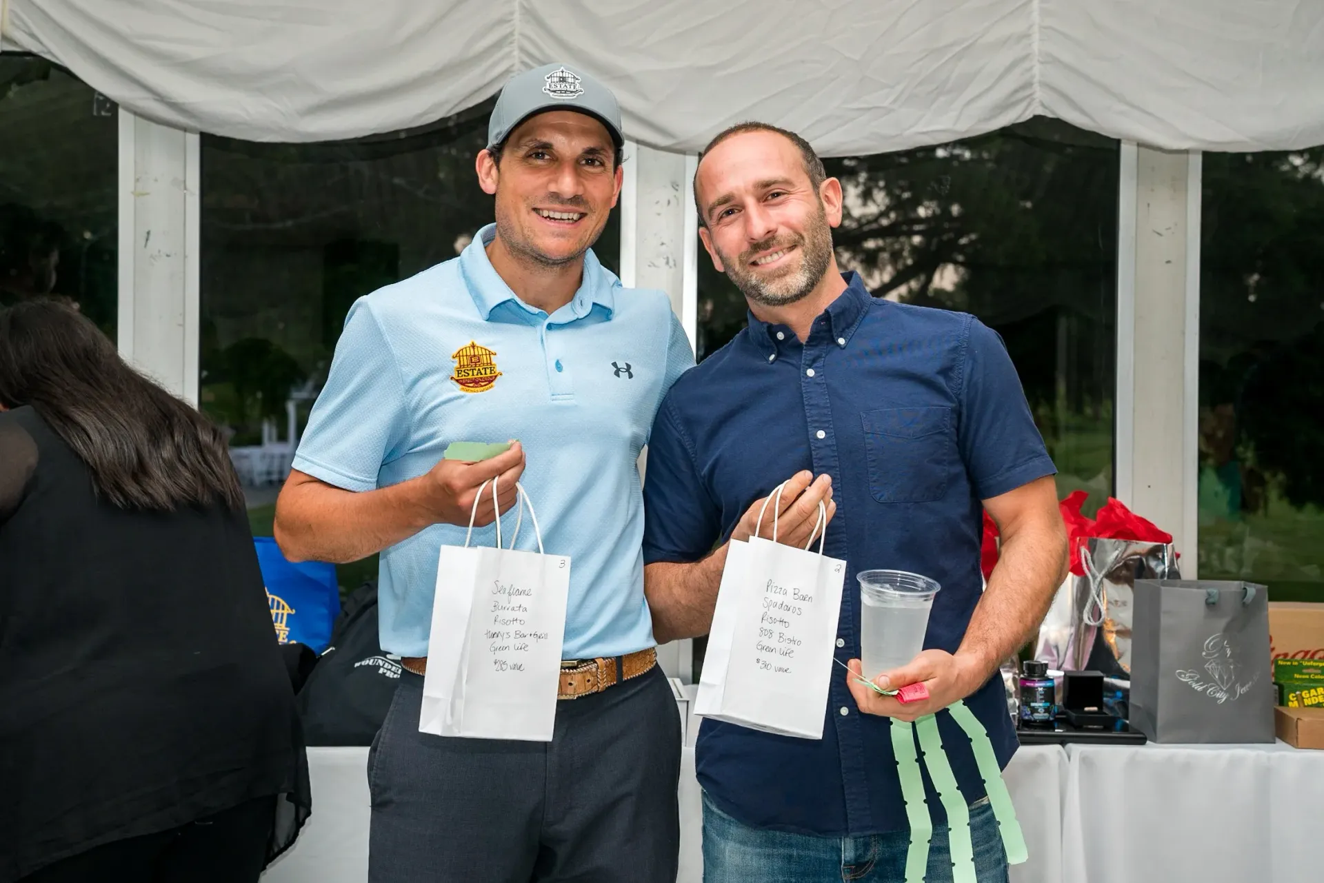 Two men are standing next to each other holding bags and drinks.