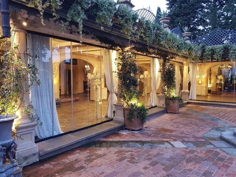 Spazio per eventi all'aperto con pareti in vetro, tende drappeggiate e piante in vaso su un patio in mattoni.