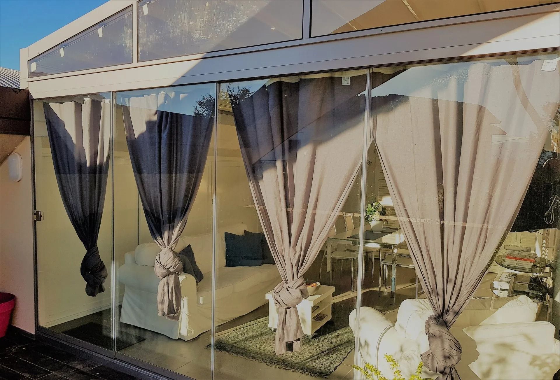Stanza con pareti in vetro e tende grigie legate. Divano bianco visibile attraverso il vetro.