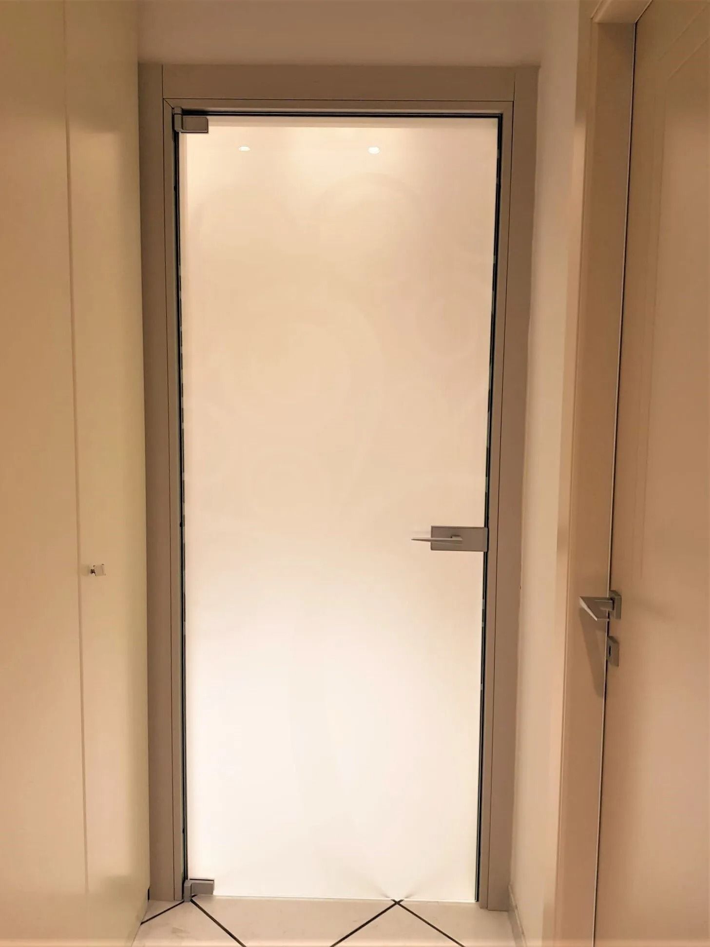 Porta in vetro smerigliato con maniglia argentata, inserita in un corridoio bianco tra due pareti.