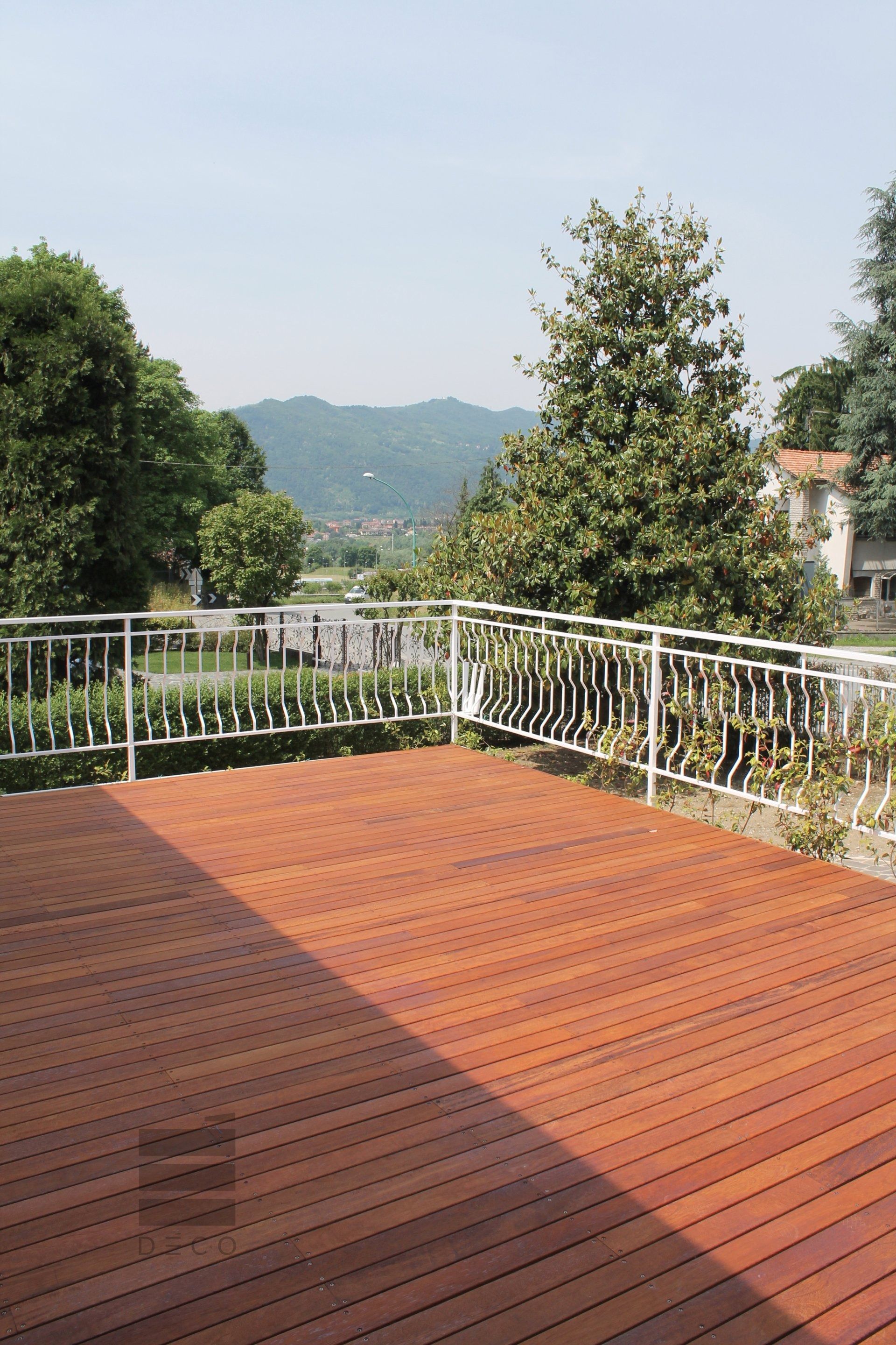 Terrazza in legno con ringhiera bianca, con vista sugli alberi e sulle montagne in lontananza sotto un cielo azzurro.