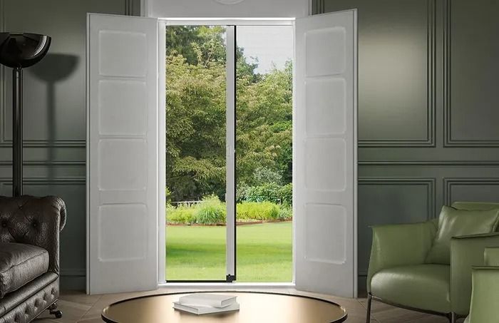 Porte bianche a due ante con zanzariera, con vista su un giardino. All'interno, una stanza con posti a sedere e un tavolino.