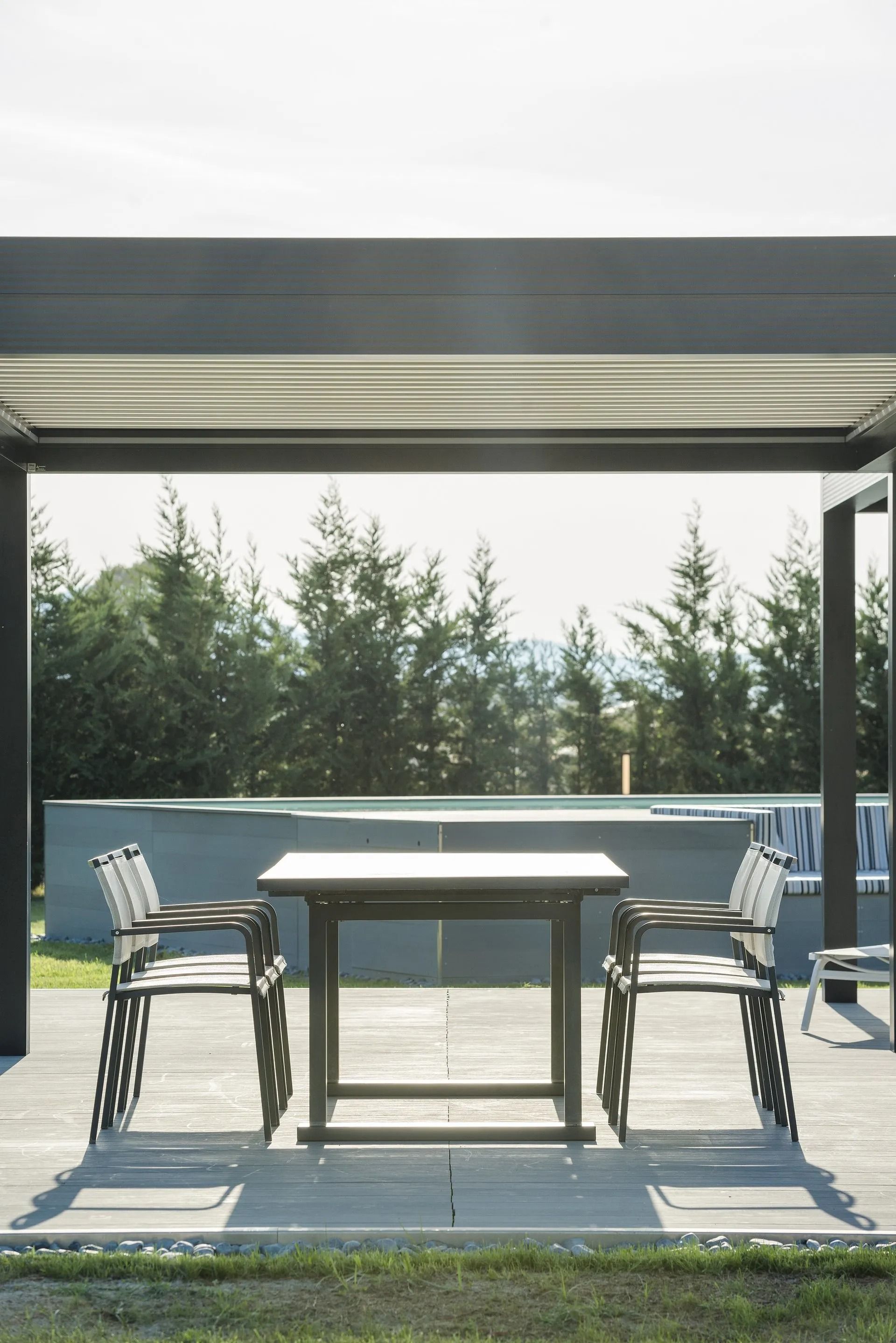 Tavolo e due sedie sotto un pergolato moderno, con vista sulla piscina e sugli alberi.