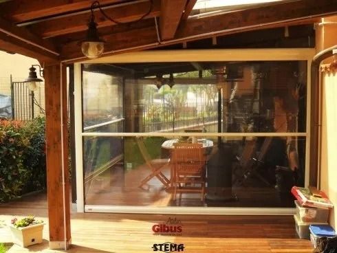 Patio chiuso con schermi trasparenti in vinile, struttura in legno e mobili da esterno visibili all'interno.
