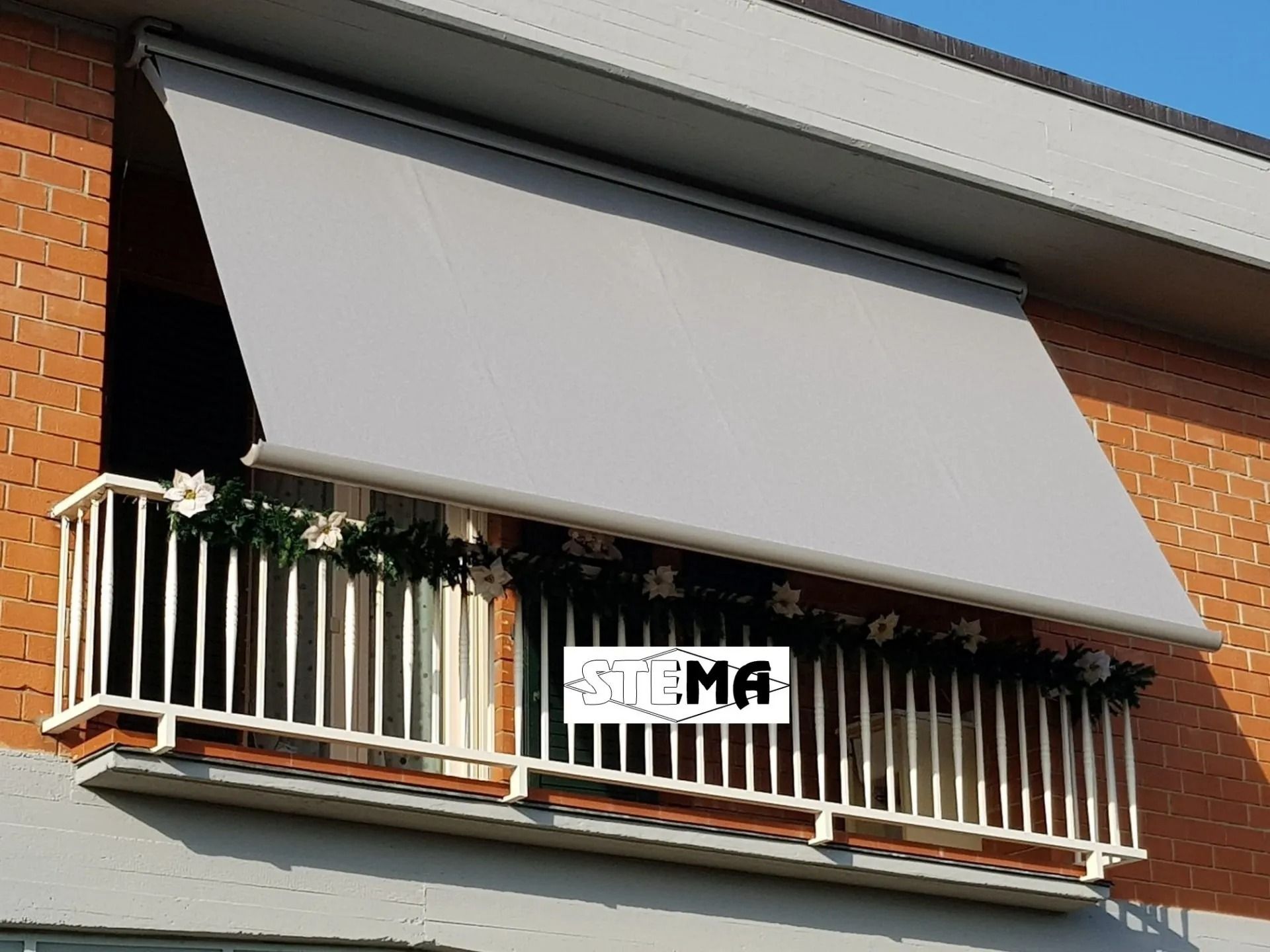 Tenda da sole retrattile grigia su un balcone con ringhiera bianca, fiori e muro di mattoni.