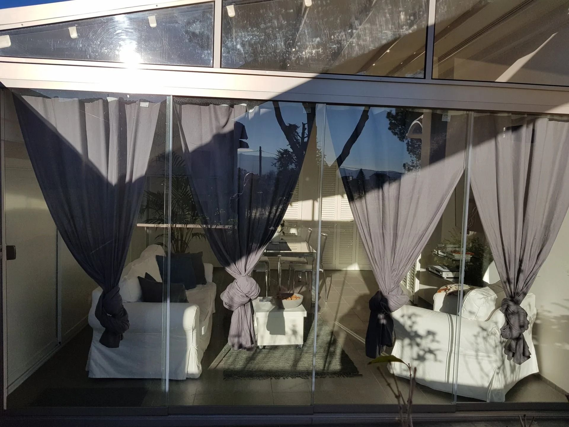 Stanza con pareti in vetro, mobili bianchi, tende grigie e luce solare.