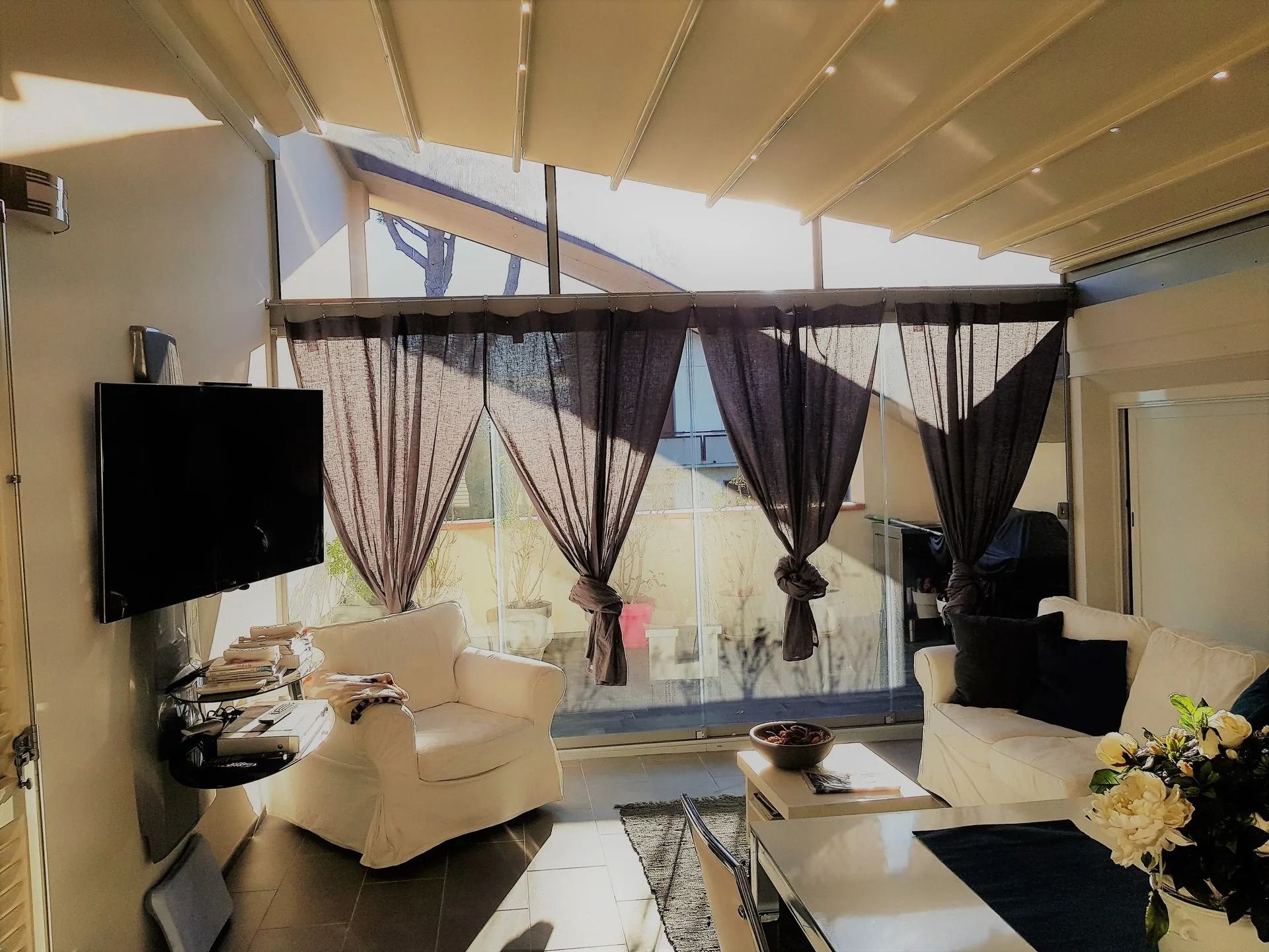 Veranda con mobili bianchi, tende scure e una TV montata su una parete.