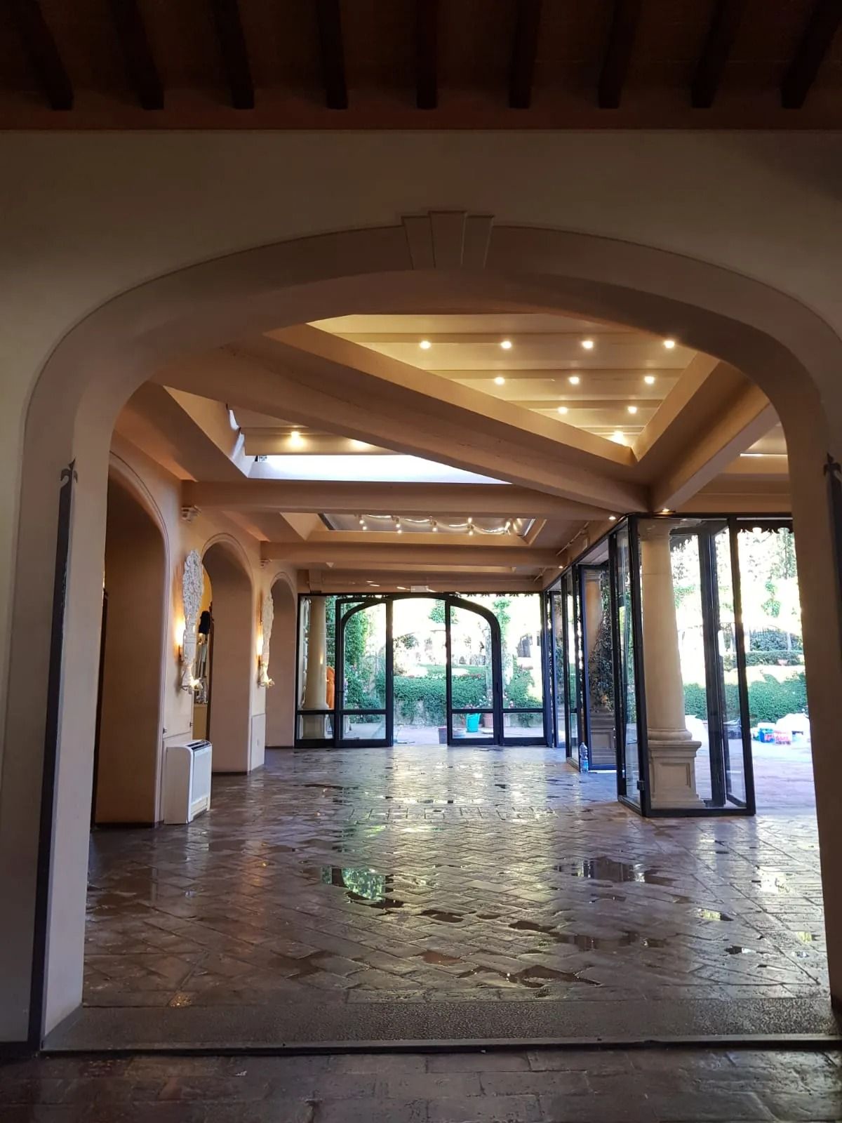 Vista interna di una stanza con porte ad arco, pavimenti in pietra e porte in vetro che si aprono su un giardino.