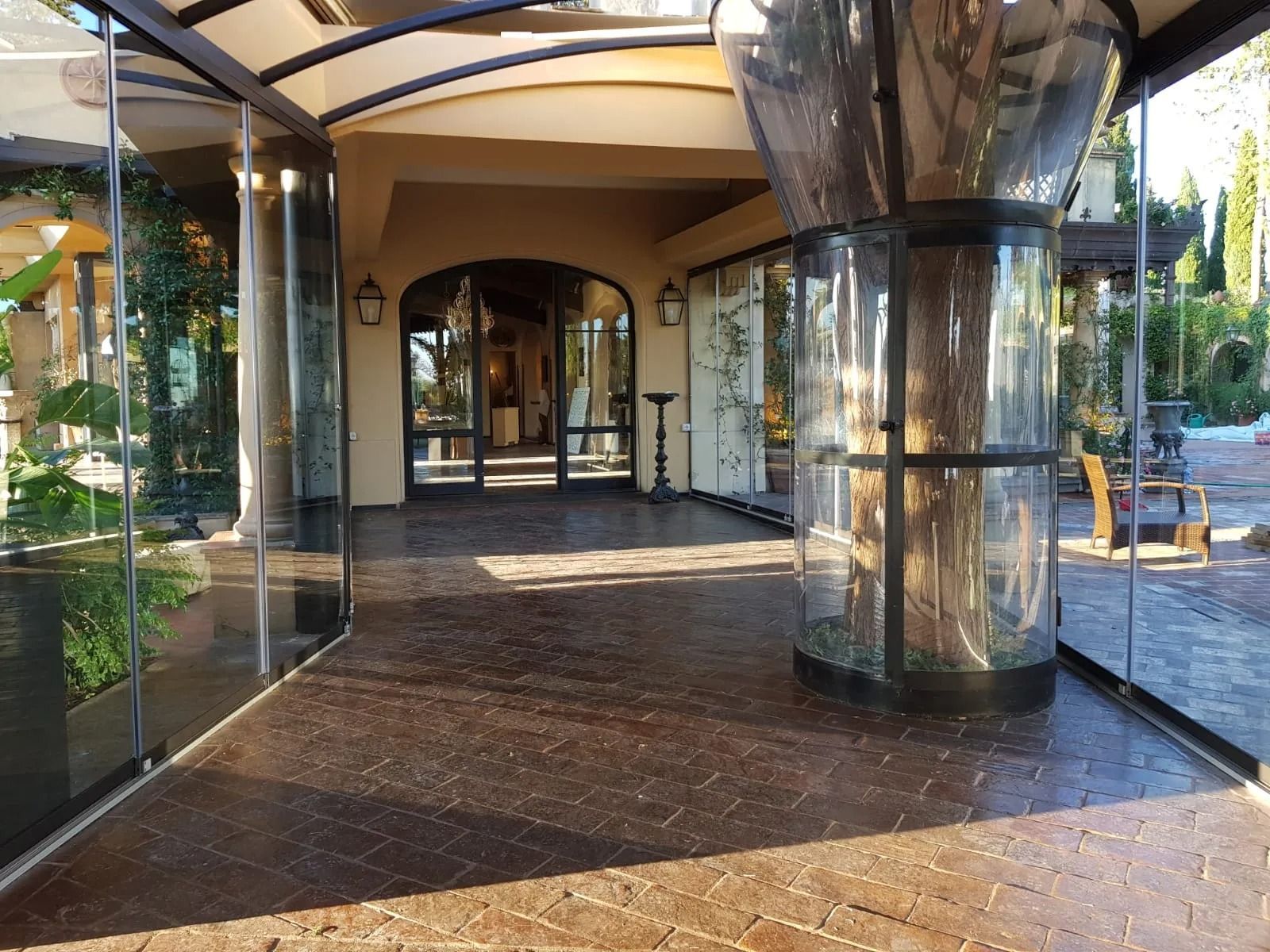 Passaggio in vetro chiuso con pavimento in mattoni testurizzati che conduce a una porta ad arco.