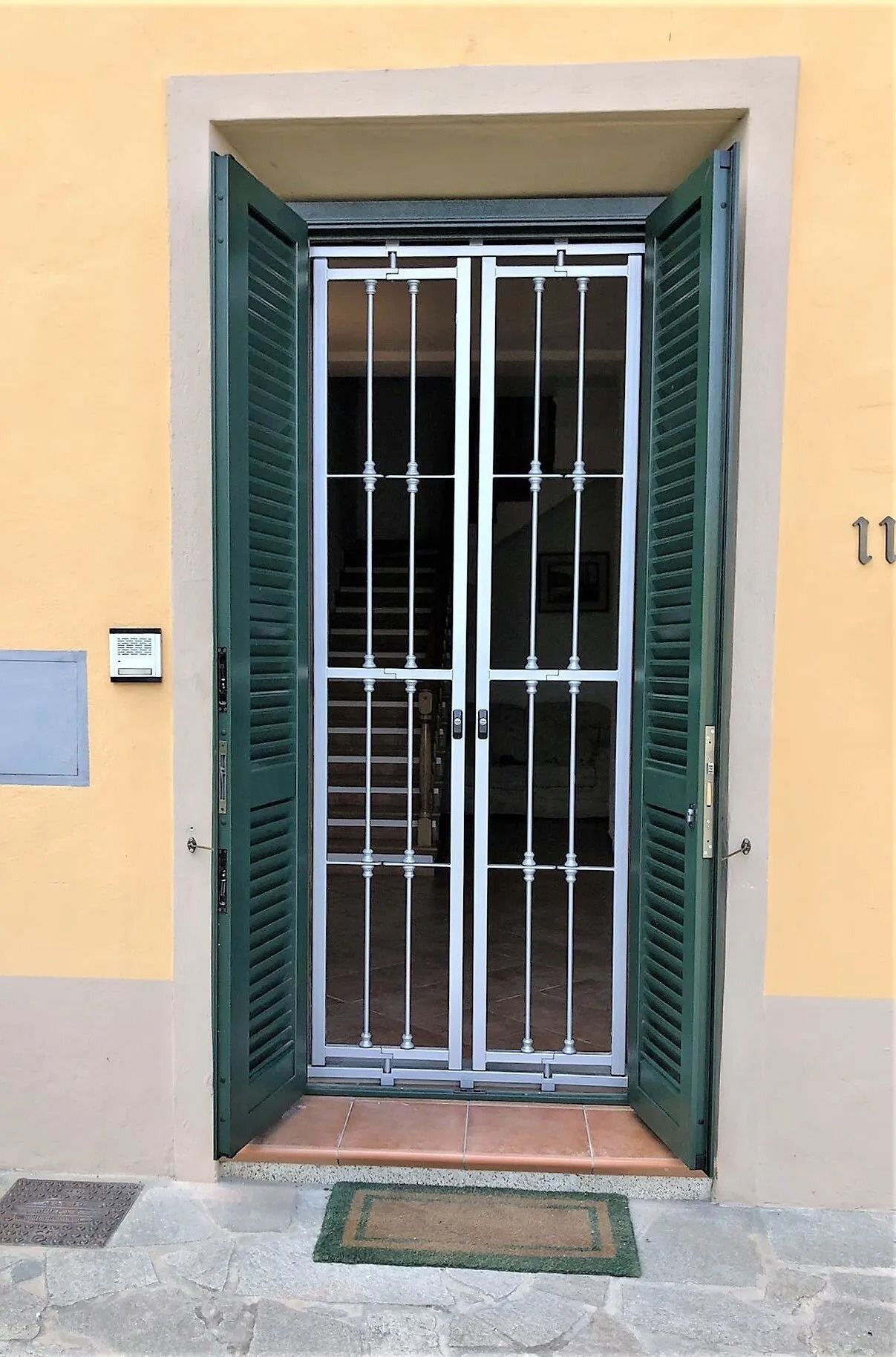 Le persiane verdi incorniciano una porta con un cancelletto di sicurezza, una soglia in mattoni e uno zerbino.