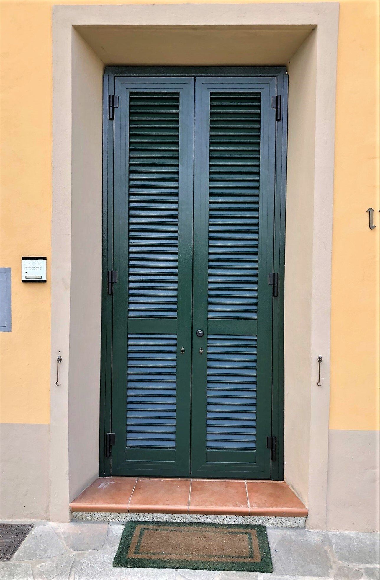 Doppie porte verdi con persiana inserite in un ingresso color marrone chiaro, con un piccolo zerbino.