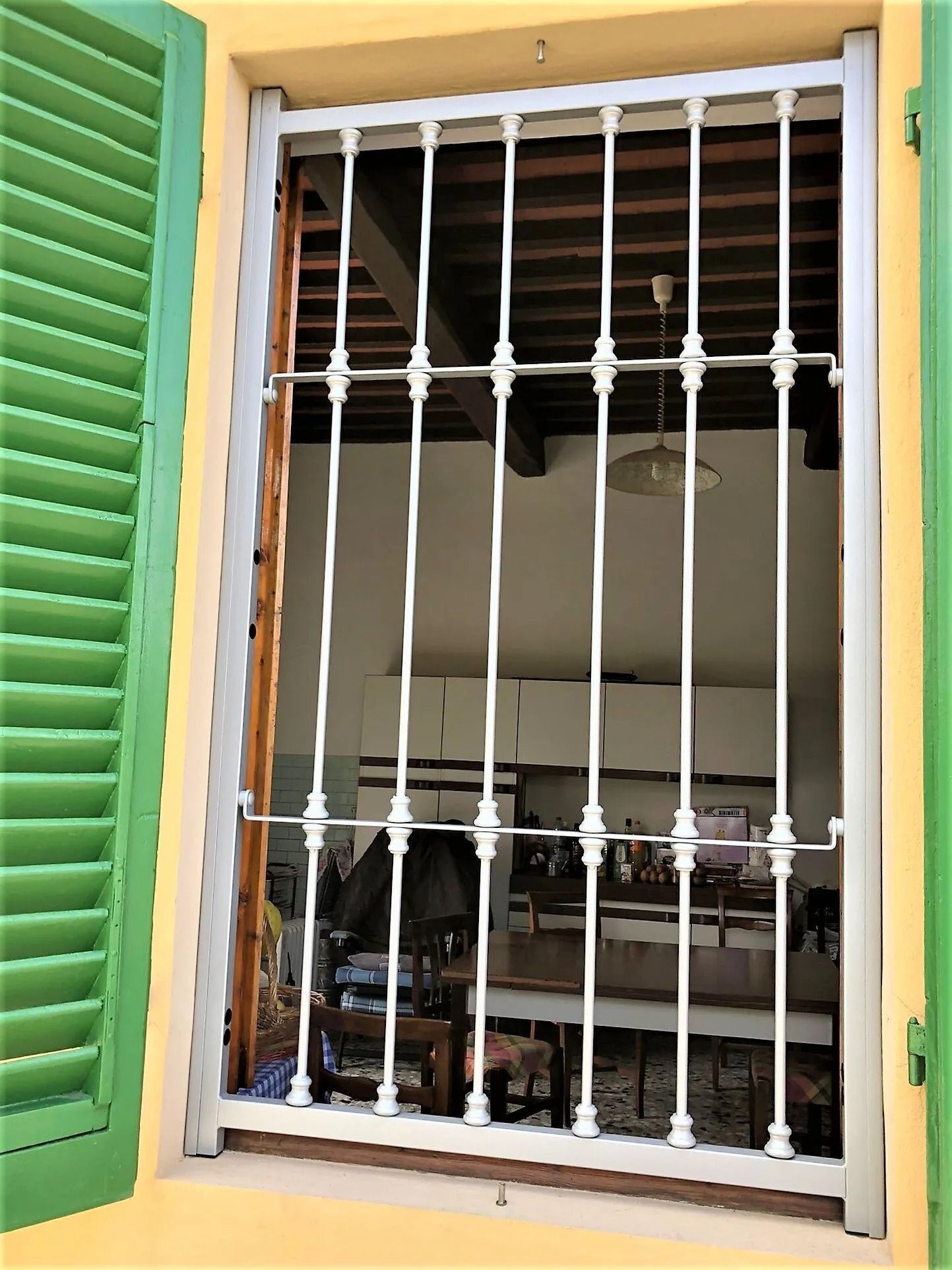 Finestra con persiane verdi, inferriate di sicurezza bianche, interno visibile: armadi, mobili e lampada a sospensione.