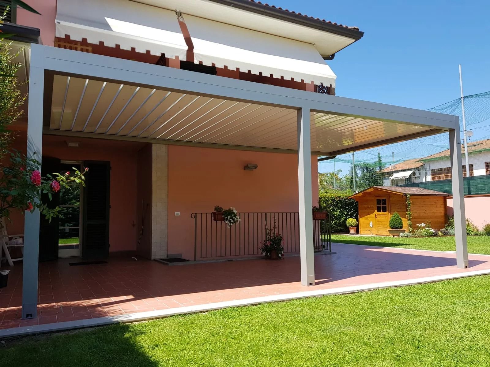 Pergola moderna in alluminio annessa a una casa rosa con prato verde.