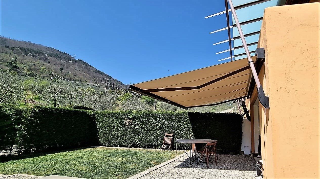 Tenda da sole marrone sopra il patio esterno con siepe, montagne e cielo azzurro.