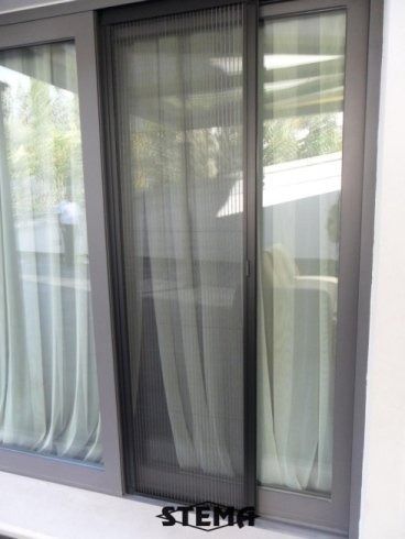 Porta scorrevole in vetro con zanzariera retrattile, telaio grigio e tende bianche.
