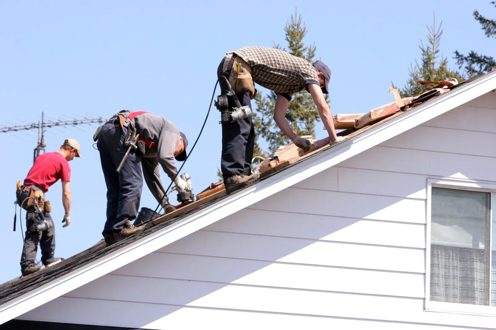 Three roofers working on a residential roof, wearing safety gear in sunny conditions.