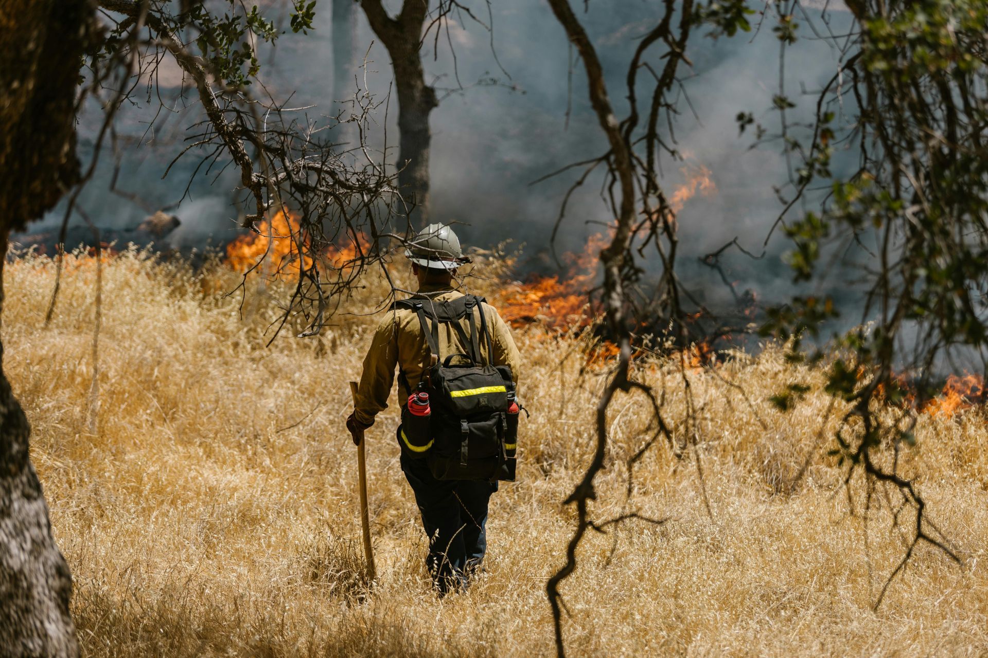 A firefighter is walking through a field with a fire in the background.