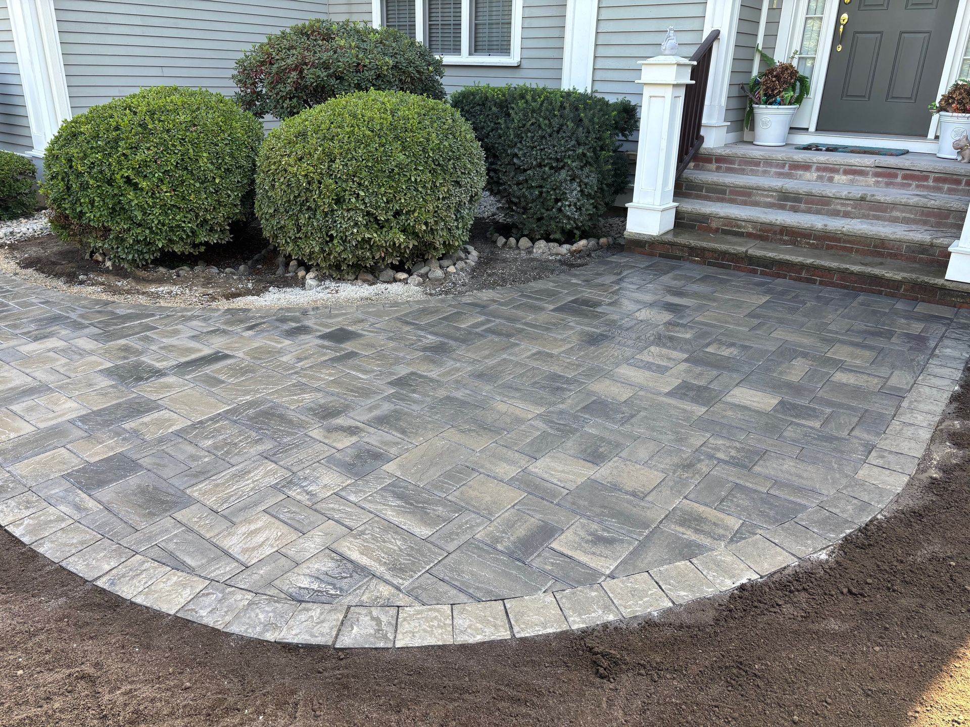 Brick patio in front of a house, surrounded by bushes and dirt.
