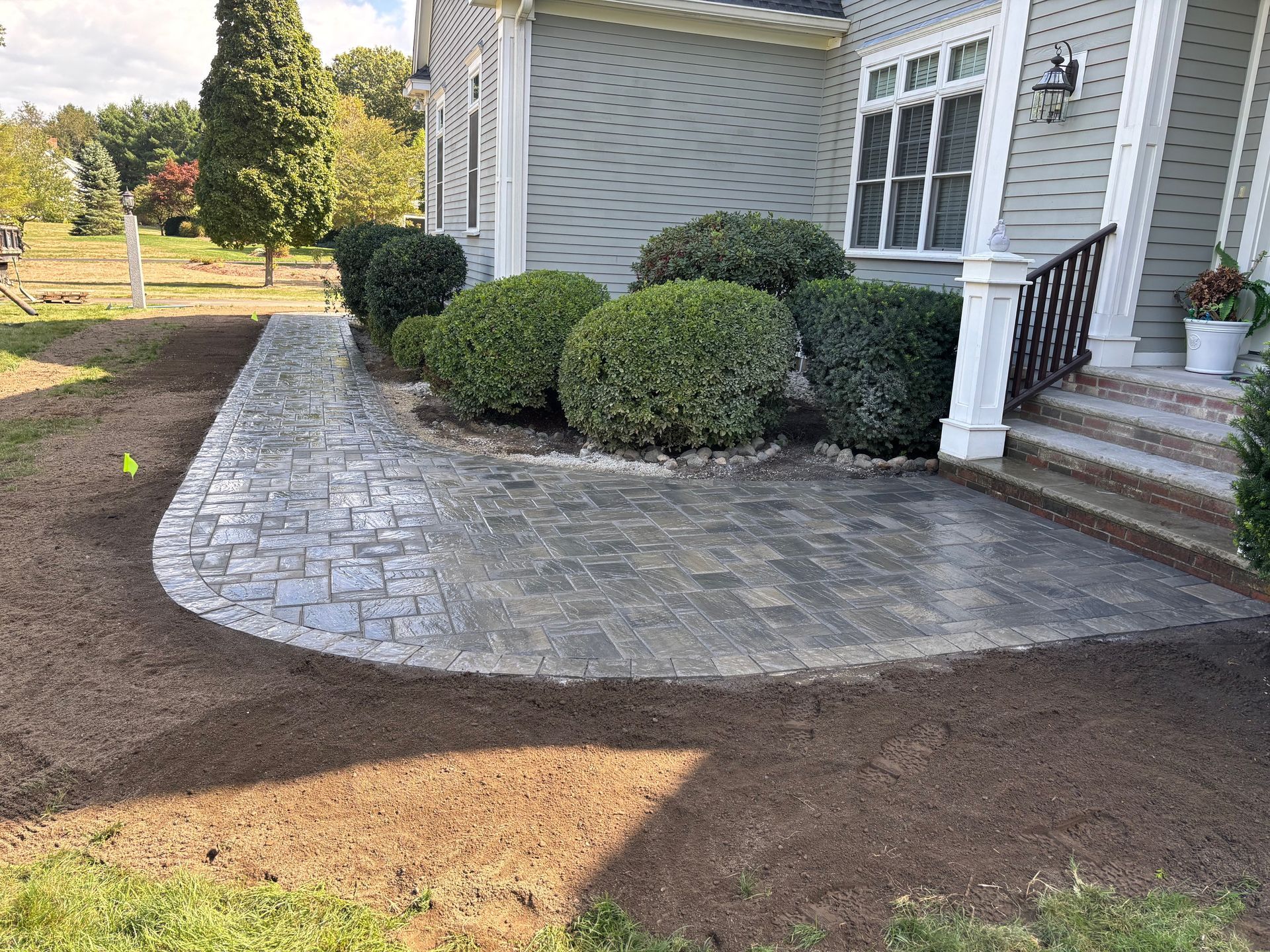 Curved brick patio in front of a gray house with green bushes and a small front porch.