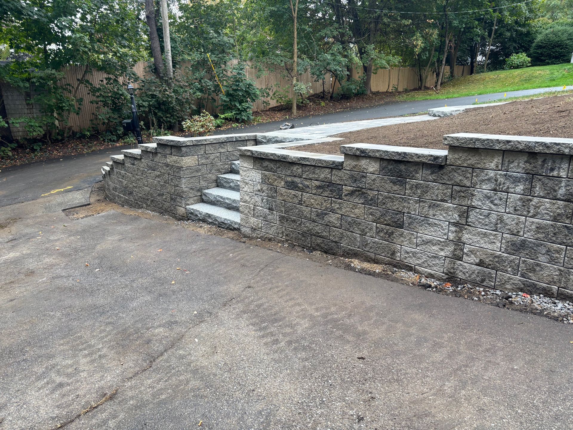Stone retaining wall with steps, separating a paved driveway from a higher area with mulch and a path.