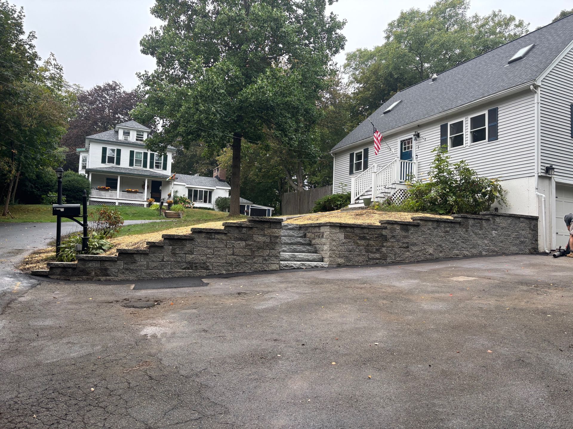 Gray stone retaining wall with steps leading to a house, other houses and trees in the background.